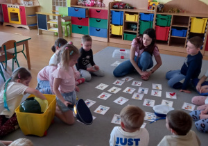 Przedszkolaki siedzą w kręgu dookała kolorowych obrazków ułożonych na dywanie.