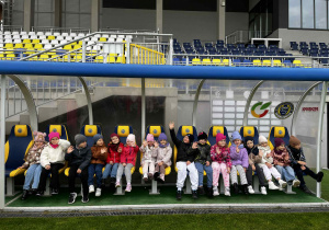 Przedszkolaki siedzą na ławce rezerwowych na boisku sportowym.