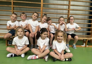 Przedszkolaki siedzą na ławeczce w hali sportowej.