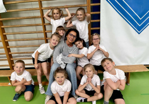 Nauczyciel z przedszkolakami na hali sportowej siedzi na ławeczce.