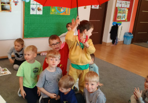 Przedszkolaki siedzą na dywanie pod czerwonym parasolem.