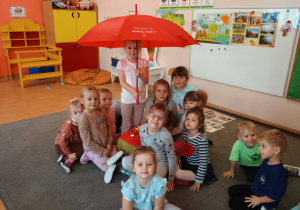 Małe dzieci siedzą na dywanie pod czerwonym parasolem.