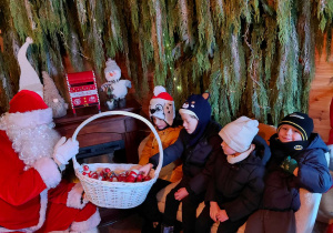 Mikołaj trzyma w ręku koszyk ze słodyczami i częstuje dzieci.